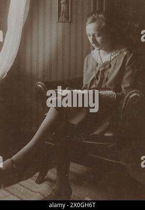 Jeune femme d'environ 25 ans assise debout dans un fauteuil en cuir rembourré avec les jambes croisées et lit un livre. Elle porte une robe de couleur claire avec un col blanc ouvert et un quilting blanc à la fin des manches et un long collier à la mode. Elle est assise sur le côté d'une fenêtre avec des rideaux blancs, d'où vous pouvez voir la lumière entrer. Sur le côté se trouve un papier peint avec des rayures verticales claires et sombres et une petite image encadrée d'une figure de la Vierge Marie, peut-être l'Uta de la cathédrale de Naumburg [traduction automatique] Banque D'Images