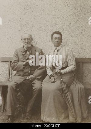 Piskotschek, inspecteur principal en bâtiment de 70 ans, avec Irma, sa fille de 50 ans, assise sur un banc en bois devant un mur de maison blanche. Homme aux cheveux gris avec une barbe blanche pleine portant un costume, fille portant une jupe pleine longueur et chemisier rayé avec patte de boutonnage couverte, contrairement à son père assis dans une posture très tordue, tous deux avec des parapluies dans leurs mains [traduction automatique] Banque D'Images