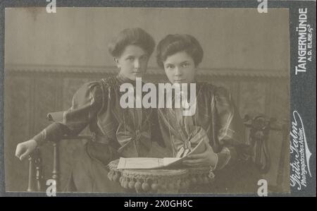 Portrait de deux soeurs jumelles (environ 17 ans) dans leur salon à la maison, assises sur des chaises avec dos lyre, devant elles un livret ouvert (programme) sur une table avec une couverture à cordon les deux filles habillées de la même façon et avec la même coiffure (chignon épinglé), jupe longue et chemisier bouffant à manches bouffantes, cravate soigneusement pliée, une des filles avec un grand bracelet en or lourd [traduction automatique] Banque D'Images