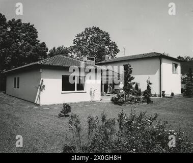 Vue d'un bungalow en forme de fer à cheval avec une extension latérale d'un étage. Le bâtiment est recouvert de tuiles à un angle peu profond et se trouve dans un terrain avec une pelouse courte. Il y a de grands arbres à feuilles caduques derrière la maison. Il y a des buissons à fleurs au premier plan. Une chaise suspendue peut être vue sur la terrasse légèrement dissimulée [traduction automatique] Banque D'Images