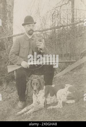 Hunter, environ 40 ans, portant un costume sombre de 3 pièces avec col rigide et cravate, assis sur un rocher avec ses jambes écartées. Il tient un fusil à double canon dans ses mains. Il porte une barbe pleine taillée et des verres en nickel. Il a mis son chapeau sur le côté avec un goût décontracté. Devant lui, dans les hautes herbes, repose son chien de chasse, probablement un chien courant allemand. Jeunes conifères en arrière-plan [traduction automatique] Banque D'Images