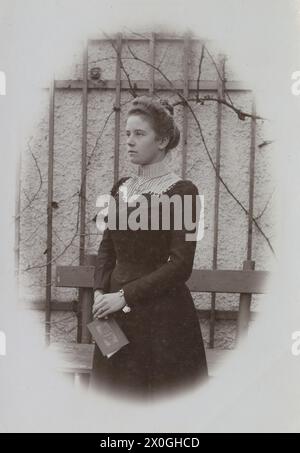 La jeune femme, âgée d'environ 20 ans, se tient devant un mur de jardin en briques avec un banc et des bordures. Elle porte une robe noire à col haut avec un corsage, un décolleté blanc et ses cheveux épinglés dans un chignon. Elle tient un mince livret dans ses mains plié devant ses genoux. Elle regarde bien au-delà de la caméra. Probablement printemps ou automne [traduction automatique] Banque D'Images