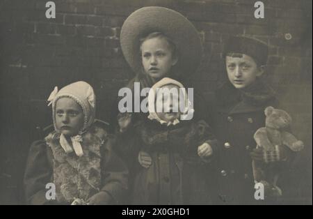 Photo de groupe de Gusti, Alfred, Oli, Toni, hiver 1909. 4 enfants âgés d'environ 2-12 ans en vêtements d'hiver. Enfants plus petits avec des casquettes blanches, fille aînée avec un grand chapeau en forme de roue porte le plus petit enfant pleurant dans ses bras, enfant à gauche avec gilet de fourrure et animal en peluche, garçon à droite avec ours en peluche et bonnet de fourrure [traduction automatique] Banque D'Images