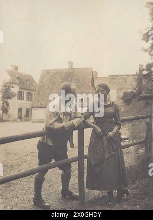 Un couple, séparé par une clôture en bois haut de la poitrine, se regarde profondément dans les yeux, penchant leurs bras contre l'échelon supérieur de la clôture. Ils sonnent leur affection l'un pour l'autre. Il y a des maisons en arrière-plan. Il porte un pantalon court en cuir, elle est une robe presque pleine longueur [traduction automatique] Banque D'Images