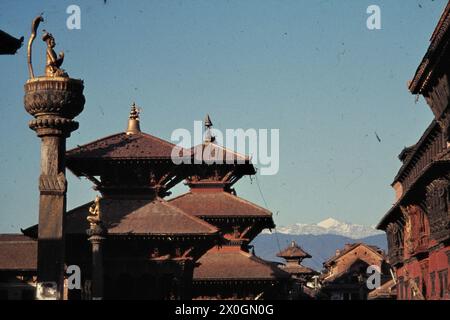 Vue sur une statue dorée de Yoga Narenda Mallas et sur les toits des pagodes et des temples du bazar de Mangal sur la place Patan Durbar avec les contreforts enneigés de l'Himalaya en arrière-plan. [traduction automatique] Banque D'Images