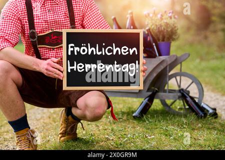 12 avril 2024 : Bavarian in lederhosen tient une pancarte avec l'inscription devant un chariot à main avec de la bière : Happy Men s Day PHOTOMONTAGE *** Bayer in Lederhose hält vor einen Bollerwagen mit Bier ein Schild mit Aufschrift : Fröhlichen Herrentag FOTOMONTAGE Banque D'Images