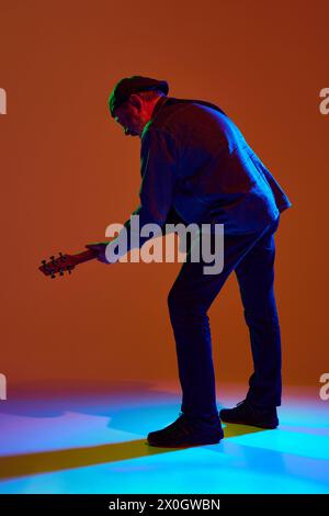 Photo vue arrière d'un musicien mature dans une tenue en denim jouant de la guitare dans une lumière néon colorée sur fond de studio orange. Banque D'Images