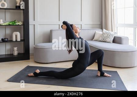 Une femme pratiquant le yoga effectue une variation de l'exercice Ashva Sanchalasana, pose cavalier, entraînement à la maison dans un sport noir sur un tapis, plein Banque D'Images