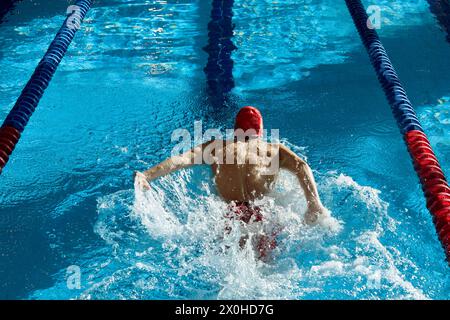Vue de dessus de musclé, athlétique jeune homme, nageur en bonnet rouge en mouvement, montrant la force, l'entraînement, la natation dans la piscine à l'intérieur Banque D'Images
