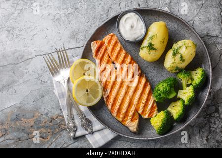 Saumon grillé diététique avec pommes de terre bouillies, brocoli, citron, herbes et sauce à la crème gros plan dans une assiette sur la table. Vue horizontale de dessus Banque D'Images