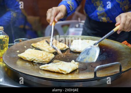 Plongez dans les rues animées de Thaïlande avec cette scène vibrante. Un vendeur qualifié prépare habilement roti croustillant, un plat de Street food apprécié Banque D'Images