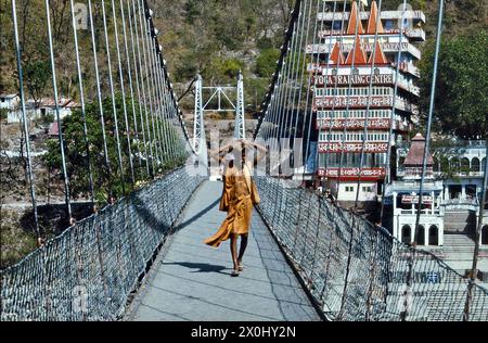 Un vieil homme, un Sadu traverse un pont suspendu à Rishikesh. Il porte des robes orange clair et des sandales. En arrière-plan, sur une colline, un bâtiment de plusieurs étages avec l'inscription : ''Yoga Training Centre''. [traduction automatique]' Banque D'Images