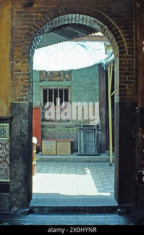 Vue par un passage de porte dans la cour intérieure d'un bâtiment résidentiel à Singapour. Les murs sont en partie couverts de briques et de tuiles vitrées. Des meubles anciens et des boîtes en bois sont installés dans la cour. Le soleil brille dans la cour. [traduction automatique] Banque D'Images