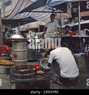 Un restaurant de rue (Hawker) à Hong Kong. Au premier plan, un homme est assis en position de squatting et se lave un pot. Un autre homme le regarde rire. Derrière lui se trouvent des tables avec parasols d'un snack-bar en bordure de route. Quelques personnes y mangent. [traduction automatique] Banque D'Images