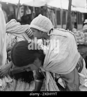 Une jeune femme porte son bébé sur son dos. L'enfant est enveloppé dans un tissu et porte un chapeau tricoté. En arrière-plan, il y a des étals du marché. [traduction automatique] Banque D'Images