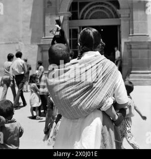 À Toluca, au Mexique, une jeune femme porte son petit enfant enveloppé dans un tissu sur son dos. Elle marche avec plusieurs autres enfants et adultes vers l'entrée d'une église sur le marché. Devant l'entrée se trouve une figure de Jésus avec une croix. [traduction automatique] Banque D'Images