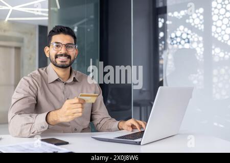 Un jeune homme joyeux dans une chemise intelligente faisant une transaction en ligne avec une carte de crédit sur un ordinateur portable dans un cadre de bureau moderne. Banque D'Images