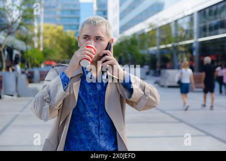 Attrayant jeune homme latin dans des vêtements formels, porte une veste et une chemise bleue, est debout sur appel parlant au téléphone à l'extérieur et buvant du café à l'air d'un Banque D'Images