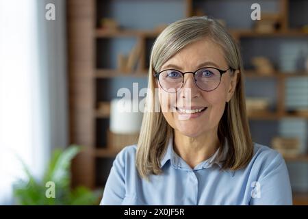 Portrait d'une femme âgée souriante avec des lunettes, portant une blouse bleue, dans un environnement de bureau moderne. Cette image capture sa confiance et son professionnalisme. Banque D'Images