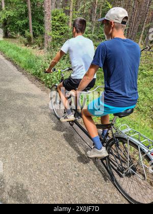 Deux adolescents en tandem sur une route étroite à travers une forêt verdoyante dans la station de vacances de Prerow, Fischland Darß, Mecklembourg-Poméranie occidentale, Allemagne Banque D'Images