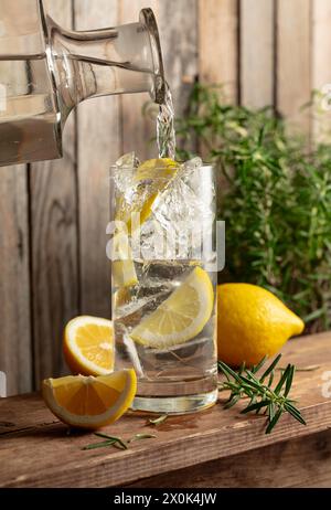 Boisson rafraîchissante d'été avec de la glace, du romarin et des tranches de citron sur une vieille table en bois. L'eau est versée d'une carafe dans un verre avec de la glace et du citron sli Banque D'Images