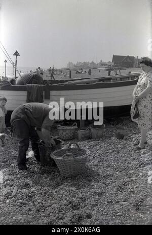 Années 1950, historique, debout dehors sur une plage de pierres, une dame regardant un pêcheur dans des échassiers avec un panier en osier triant sa prise, Angleterre, Royaume-Uni. Banque D'Images