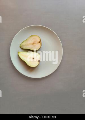 Une poire coupée en deux avec tige repose avec les deux moitiés sur une plaque gris clair, thème nutrition naturelle saine avec des fruits Banque D'Images