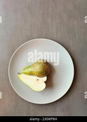 Une poire coupée en deux avec tige repose avec les deux moitiés sur une plaque gris clair, thème nutrition naturelle saine avec des fruits Banque D'Images