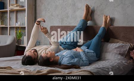 Caucasien couple heureux couché à l'envers ensemble sur le lit de confort se détendre avec le téléphone mobile défilant réseau social achetant en ligne des achats sur Internet Banque D'Images