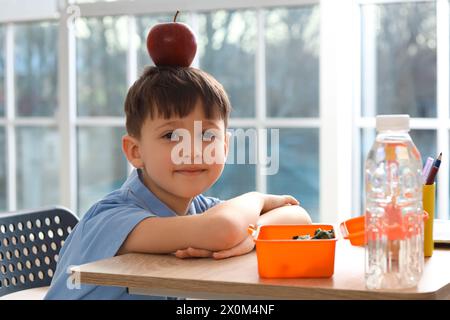 Mignon petit écolier avec pomme sur la tête pendant le déjeuner en classe Banque D'Images