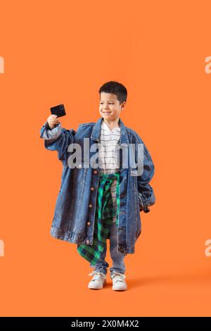 Mignon petit garçon asiatique dans des vêtements adultes avec téléphone portable prenant selfie sur fond orange Banque D'Images