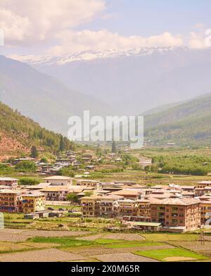 Une vue sur les champs, le logement et Paro Chhu (rivière) dans la ville de Paro dans la vallée de Paro avec des montagnes avec de la neige au sommet au printemps, l'ouest du Bhoutan. Banque D'Images