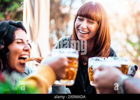 Un groupe d'amis partage un joyeux toast avec des tasses à bière, tout en se livrant à une délicieuse pizza, - la lumière du soleil filtrant à travers les arbres. Banque D'Images