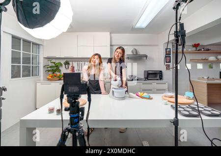 Deux femmes asiatiques dans un tablier, souriant et cuisinant dans une cuisine avec appareil photo et équipement d'éclairage pour la vidéo et la séance photo, enregistrement vidéo vlog pour s. Banque D'Images