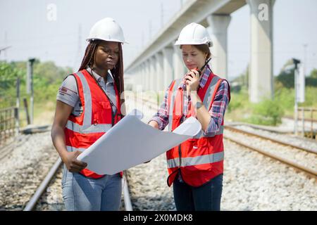 Deux ingénieurs de construction ou de construction avec casque de sécurité et gilet haute visibilité examinant les travaux à effectuer ou les progrès sur un projet de voie ferrée pendant le maintien Banque D'Images