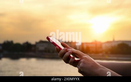 Personne féminine touchant l'écran de son téléphone avec son doigt devant le fond de la ville pendant le coucher du soleil. Banque D'Images