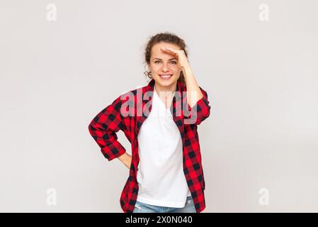 Femme portant une chemise rouge décontractée ombrant ses yeux avec la main, regardant au loin et souriant. Prise de vue en studio. Banque D'Images
