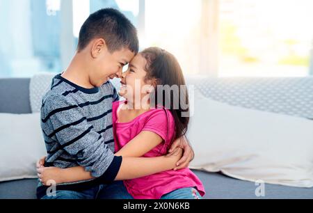 Jeunes, enfants et heureux avec câlins, sourire et joie dans la maison familiale comme les enfants dans la jeunesse. Frères et sœurs, mignons et câlins comme frère et soeur sur le canapé pour Banque D'Images