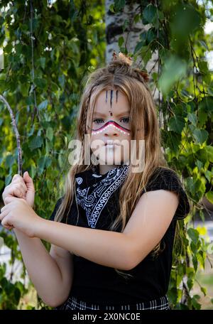 Fille de tribu avec les cheveux longs et le maquillage traditionnel habiller l'enfant de jour d'école tenant un bâton dans les bois Banque D'Images