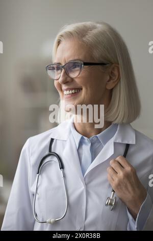 Heureuse femme médecin mûre médecin en uniforme posant à l'intérieur Banque D'Images