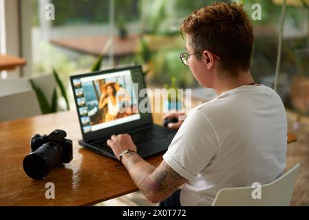 Éditeur de photo professionnel fonctionne sur ordinateur portable dans un espace de coworking lumineux. Tatoué, personne élégante édite l'image, focalisé sur l'écran, caméra à côté. Moderne Banque D'Images