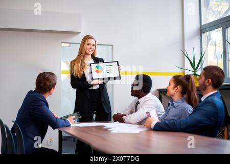 PDG féminin confiant présente le graphique coloré sur ordinateur portable convertible à l'équipe d'affaires diversifiée et multiethnique. Pro Young leader mène une séance de stratégie Banque D'Images