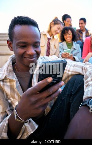 Gars noir souriant utilisant un téléphone intelligent. Homme afro-américain tenant la cellule à l'extérieur avec des amis Banque D'Images