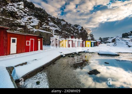 Une scène hivernale sereine capture une rangée de chalets rouges, jaunes et blancs vifs le long d'une entrée de mer partiellement gelée, avec des rochers enneigés dans le Banque D'Images