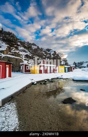 Une scène hivernale sereine capture une rangée de chalets rouges, jaunes et blancs vifs le long d'une entrée de mer partiellement gelée, avec des rochers enneigés dans le Banque D'Images