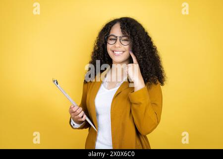 Femme d'affaires afro-américaine avec des papiers dans les mains sur fond jaune très heureuse et excitée faisant un geste gagnant avec les bras levés, souriant Banque D'Images