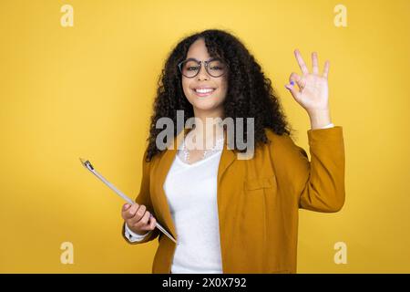 Femme d'affaires afro-américaine avec des documents dans les mains sur fond jaune faisant signe OK avec les doigts et le sourire, excellent symbole Banque D'Images