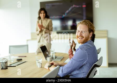 Un homme barbu sourit à la caméra tandis qu'une femme présente un graphique sur un écran dans une salle de réunion moderne. Banque D'Images