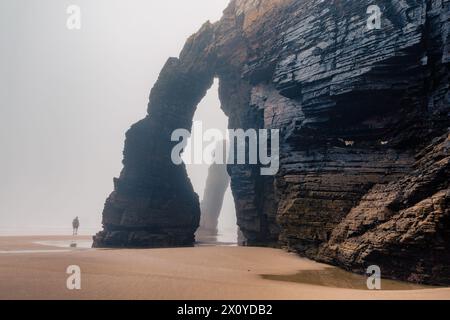 Touriste marchant le long de l'arche naturelle sur la plage des cathédrales en Galice, en Espagne. Silhouette d'homme dans le paysage brumeux avec Playa de Las Catedrales Catedrais Banque D'Images