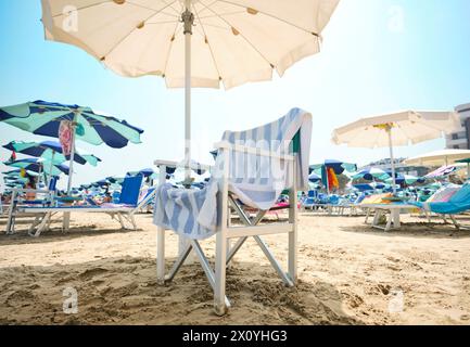 Chaise avec serviette rayée sous un parasol le à la plage sur une journée d'été ensoleillée. En arrière-plan, la plage équipée de chaises longues et parasol Banque D'Images